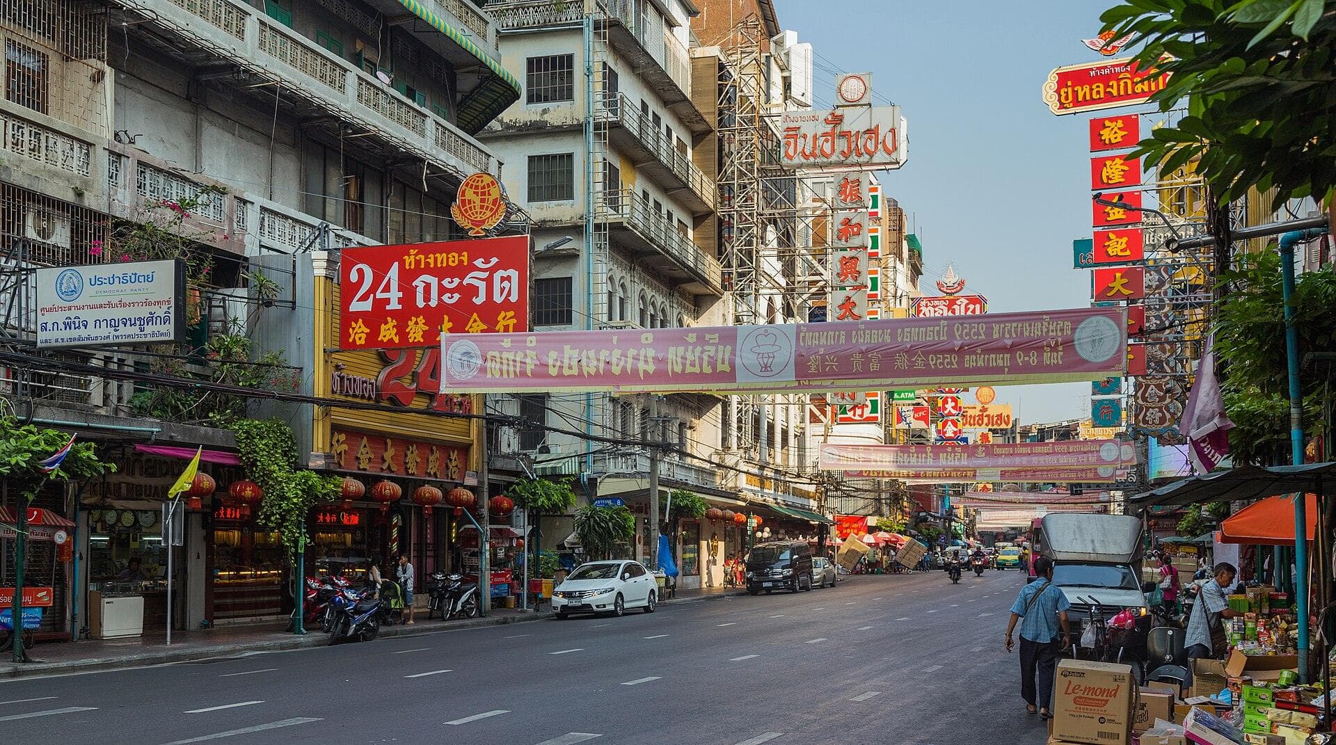 Yaowarat Road in Bangkok's Chinatown by day, lined with tall vertical shop signs in Thai and Chinese script, gold shops and shophouse terraces, with cars and motorcycles moving down the wide street.