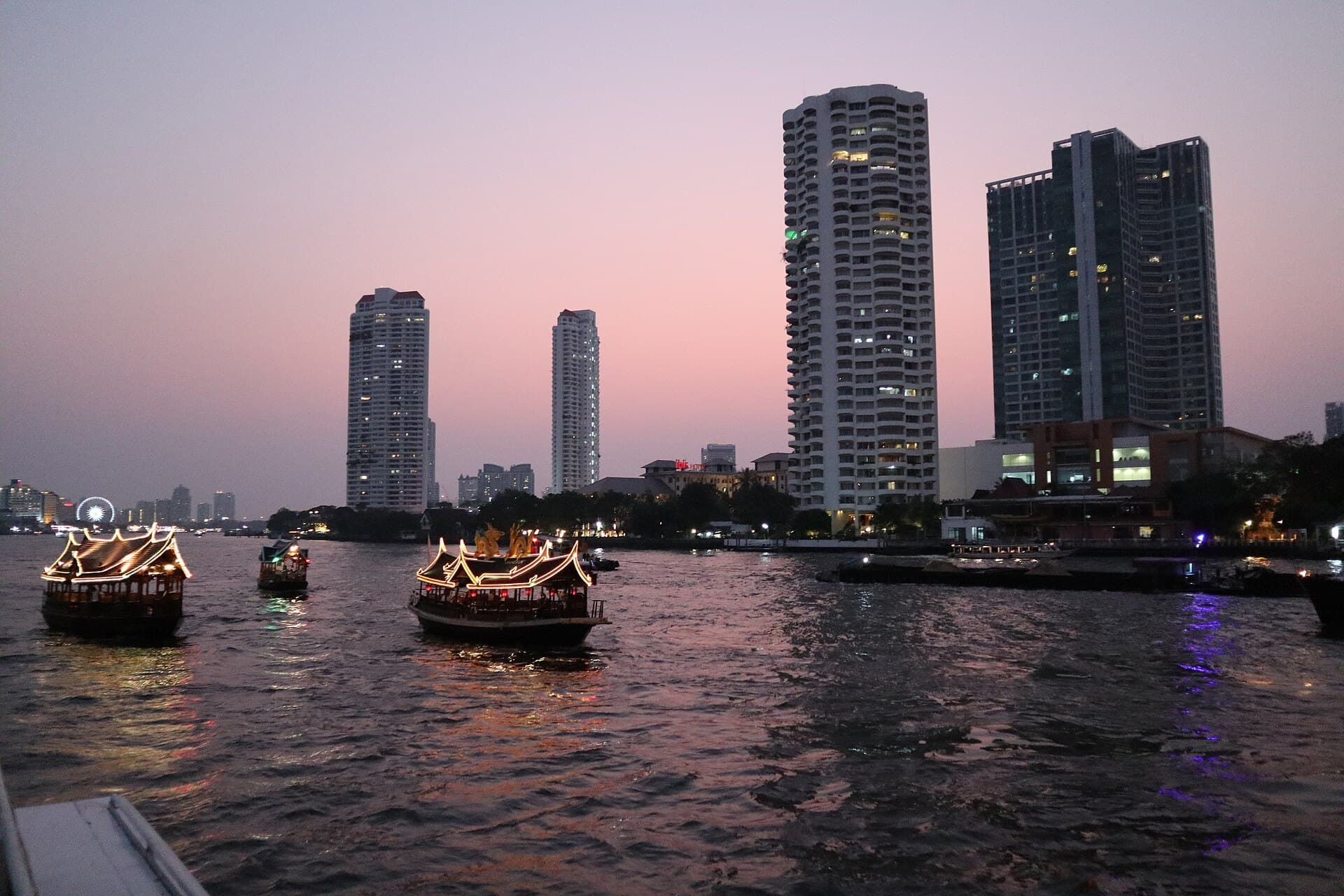 The Chao Phraya river in Bangkok just after sunset, its surface reflecting a pink and violet sky, with two lantern-lit dinner boats mid-river and riverside high-rise towers rising along the far bank.