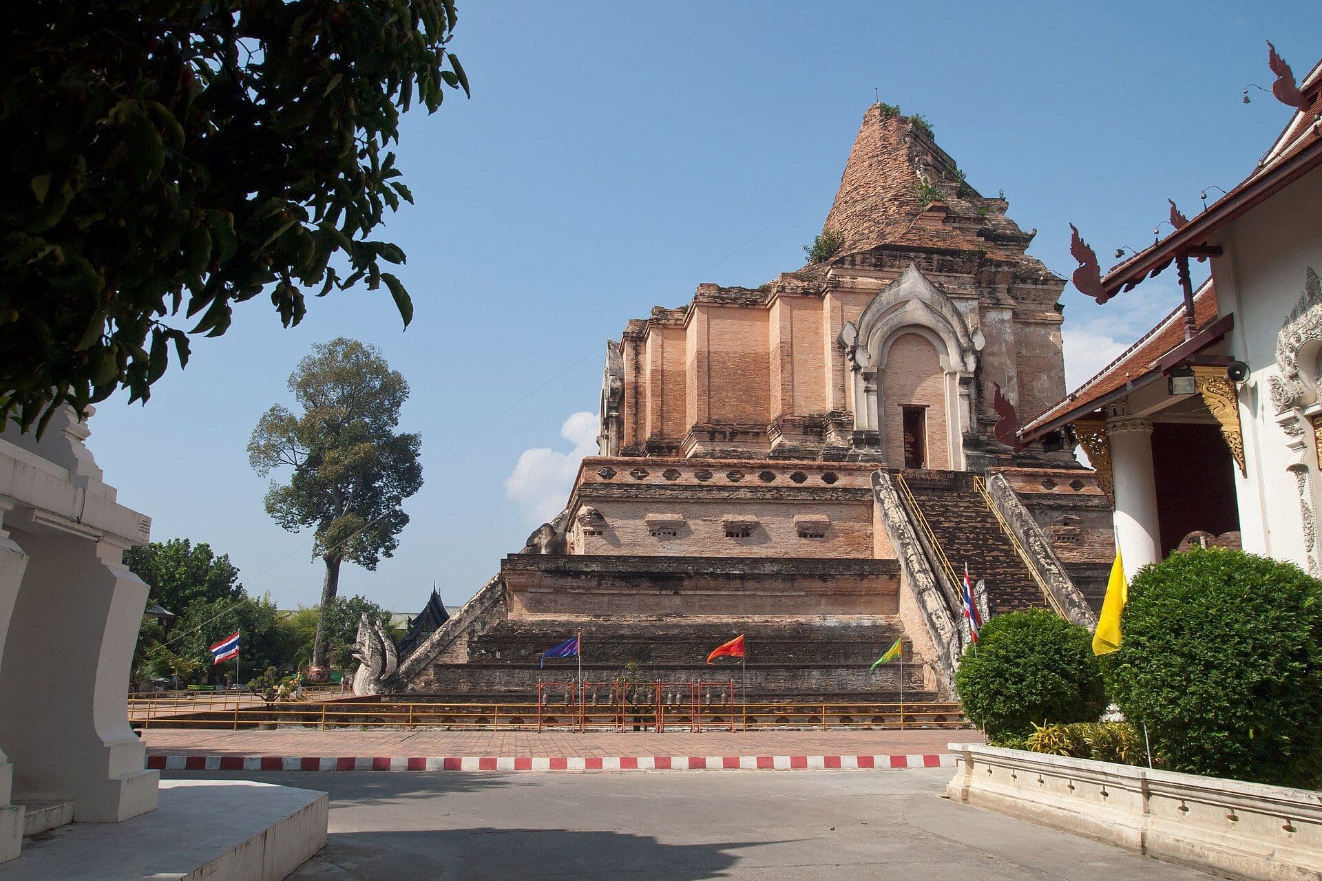 The ruined brick chedi of Wat Chedi Luang in Chiang Mai rising in stepped tiers against a clear blue sky, with a naga-flanked stairway climbing its flank, small coloured flags along the base and the white and gold eaves of the viharn at the right of frame.
