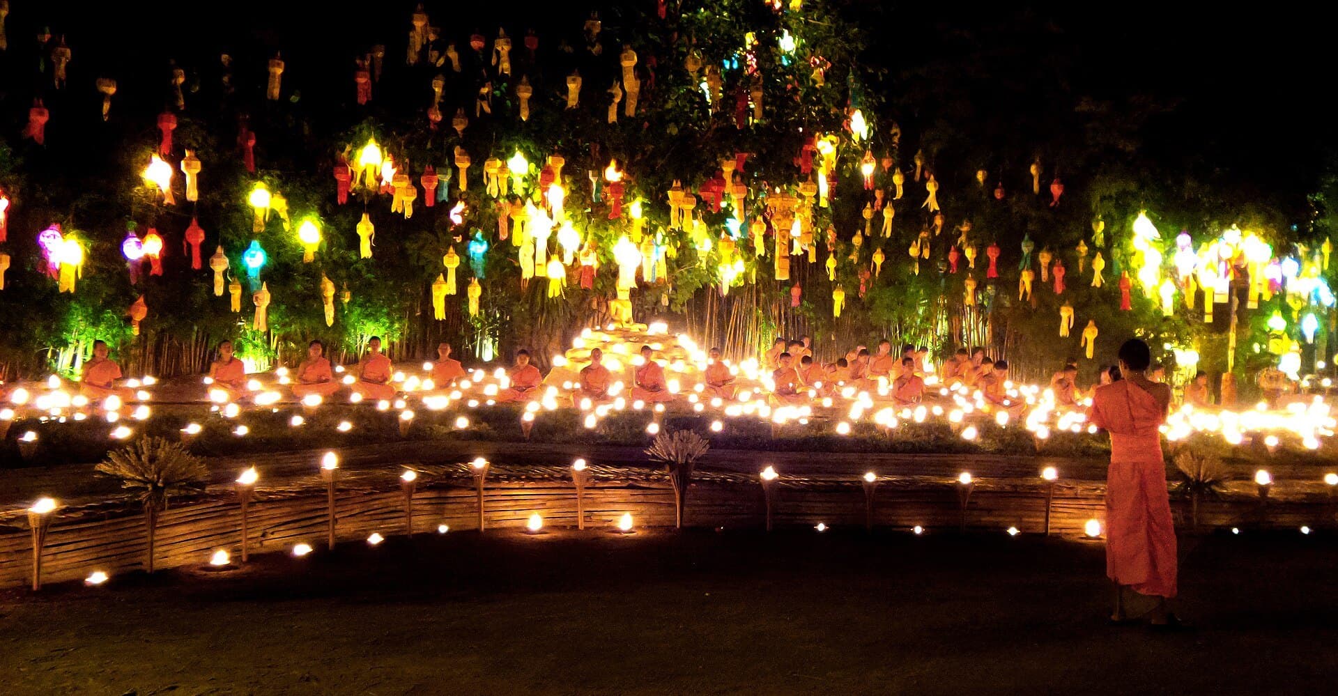 A Loi Krathong ceremony at a Chiang Mai temple after dark: rows of monks in orange robes seated among hundreds of small candle flames, beneath tiers of glowing yellow, red and blue paper lanterns strung through the trees, with one monk standing in the foreground.
