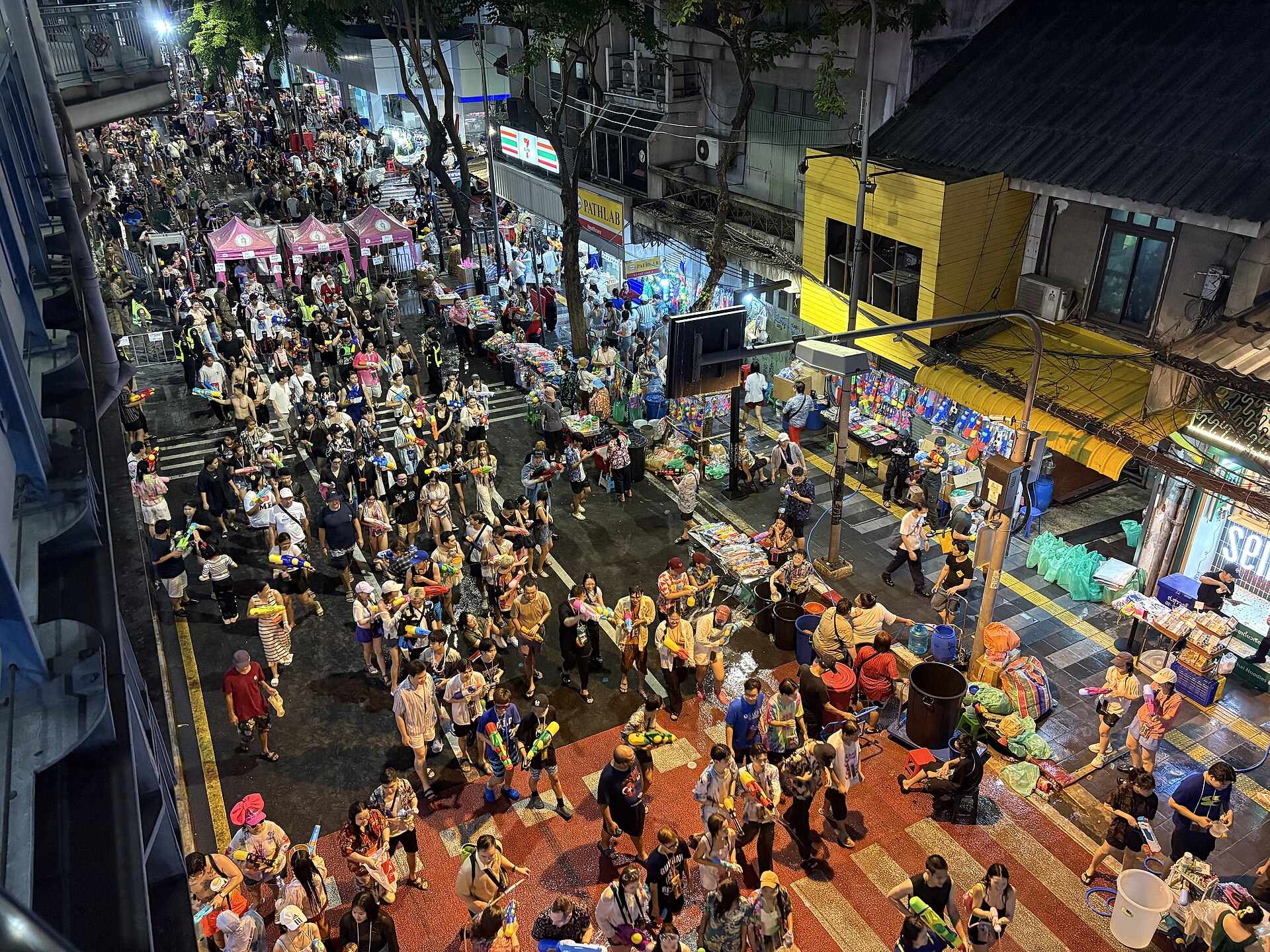 Songkran on Si Lom Road in Bangkok seen from above at night: the closed road packed with people carrying water pistols, pink-canopied stalls and pavement vendors selling more water guns, and the wet red and orange crossing markings shining under the streetlights.