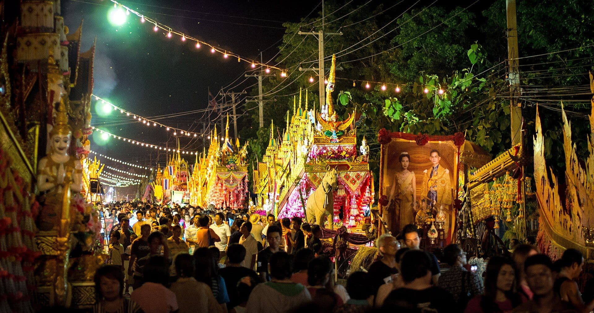The night procession at the Yasothon rocket festival in Isaan: ornate gilded ceremonial floats with tiered spires lining both sides of the street under strings of festoon lights, with a dense crowd of spectators filling the road between them.