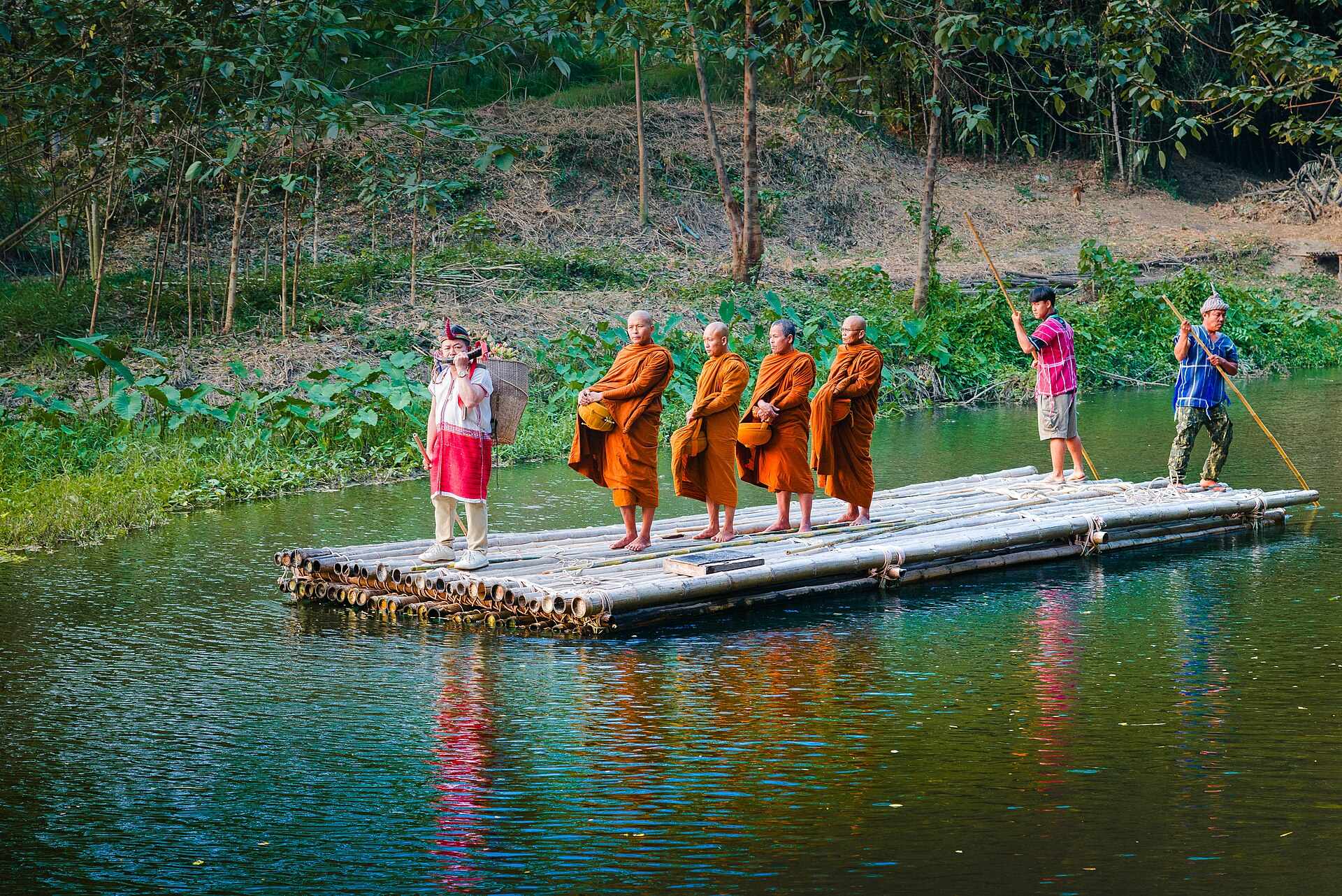 Four monks in orange robes standing with their alms bowls on a bamboo raft being poled along the green Phachi river, a woman in a red hill-tribe tunic carrying a woven back-basket beside them, with dense forest rising from the bank behind and their reflections stretching across the water.