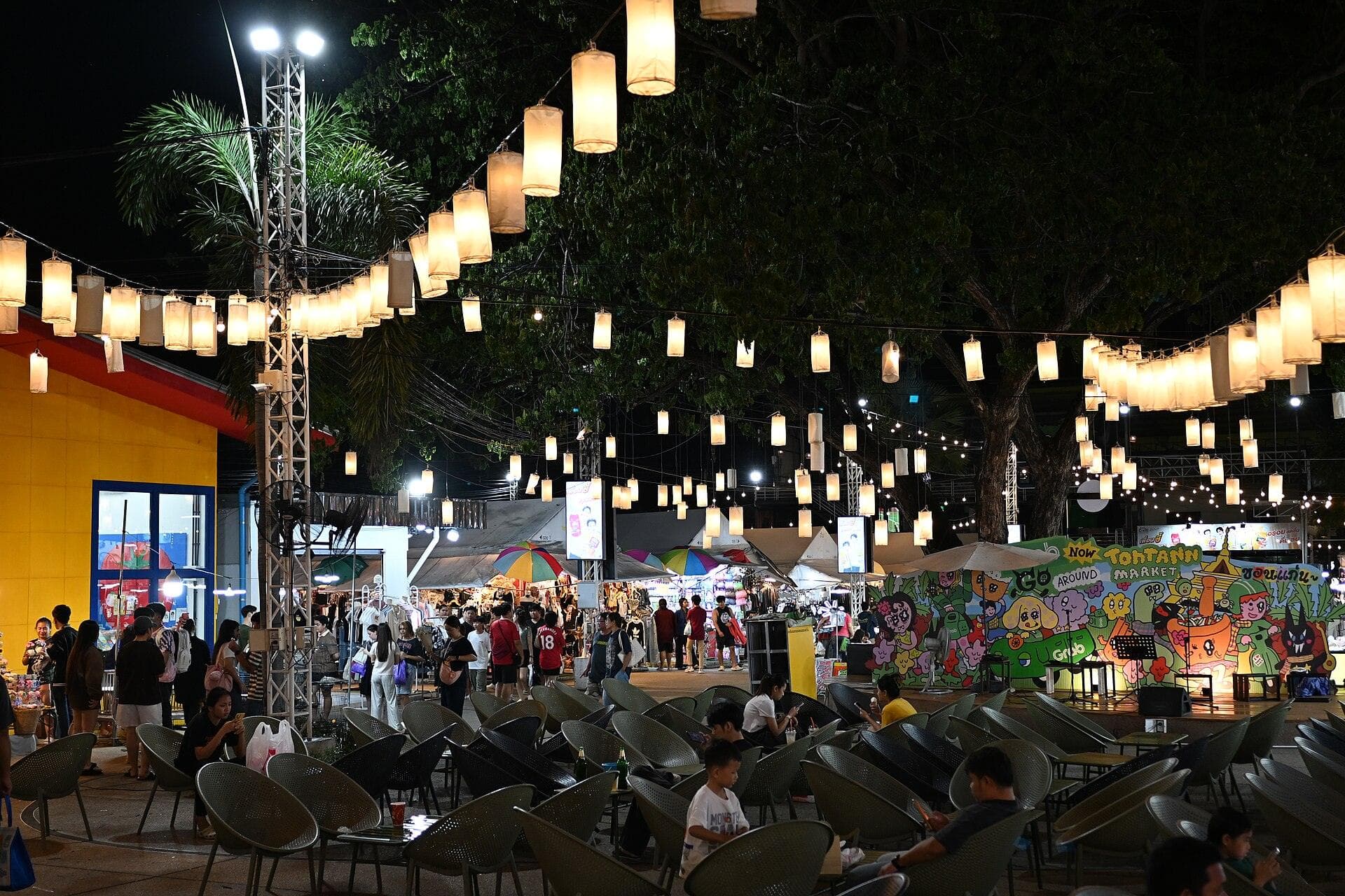 A Thai night market lit by dozens of warm paper lanterns strung in long rows overhead, with clothing and food stalls under white canopies behind, a brightly painted mural along the far wall and shoppers resting at moulded plastic chairs in the foreground.
