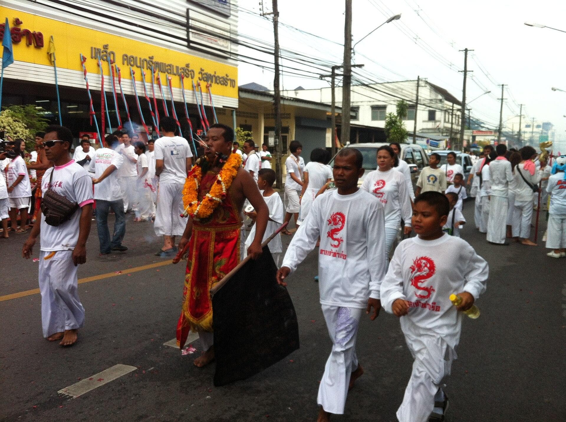 A street procession during the Phuket Vegetarian Festival: devotees dressed head to toe in white walk down the road, led by an entranced devotee in red and gold robes hung with marigold garlands who carries a black banner and has a thin skewer through his cheek, past a yellow shopfront strung with red and blue ceremonial poles.