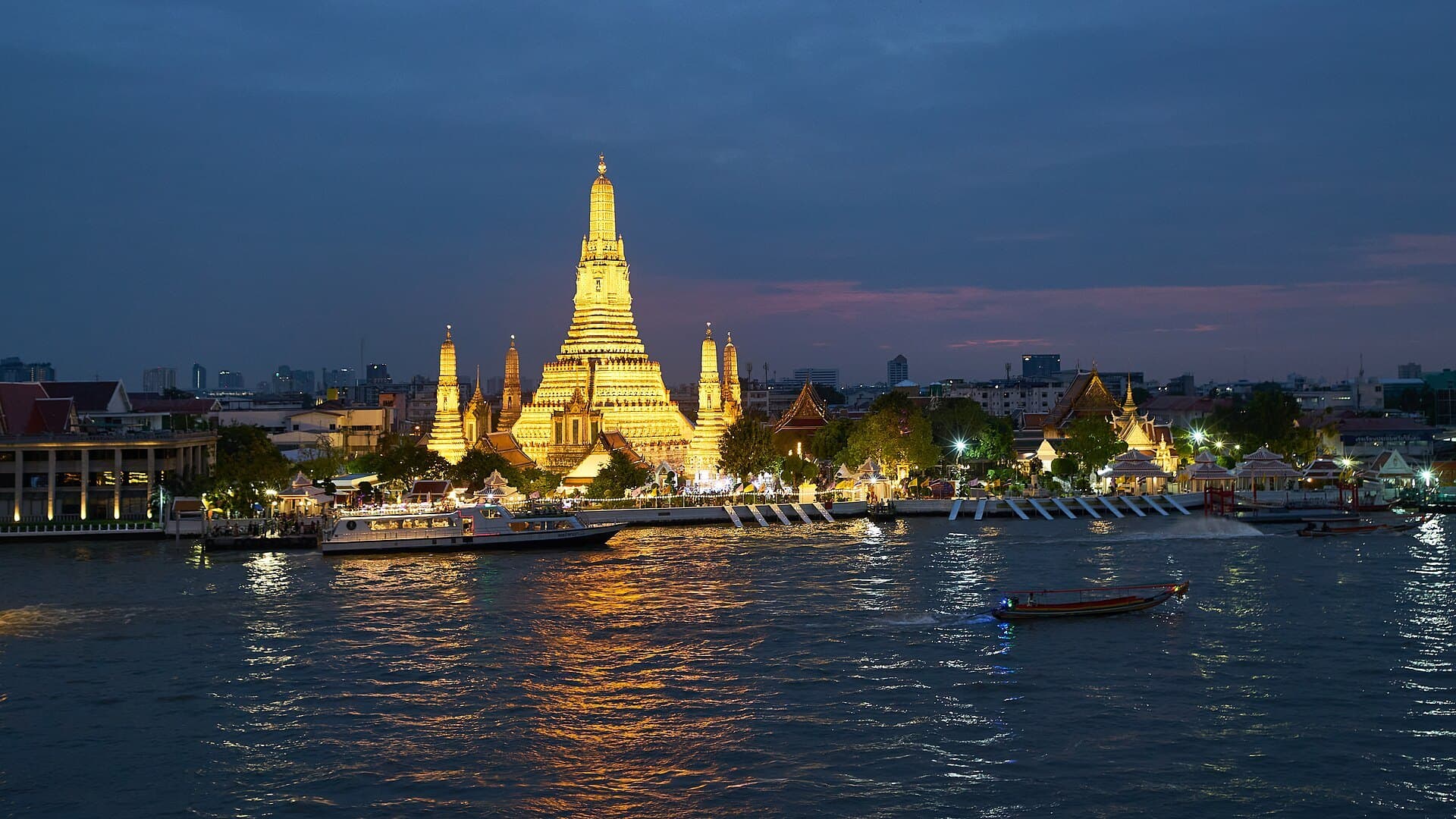 Wat Arun floodlit at dusk on the west bank of the Chao Phraya, its tiered central prang and four satellite spires glowing gold against a deep blue sky, with the temple's light doubled in the dark water below.