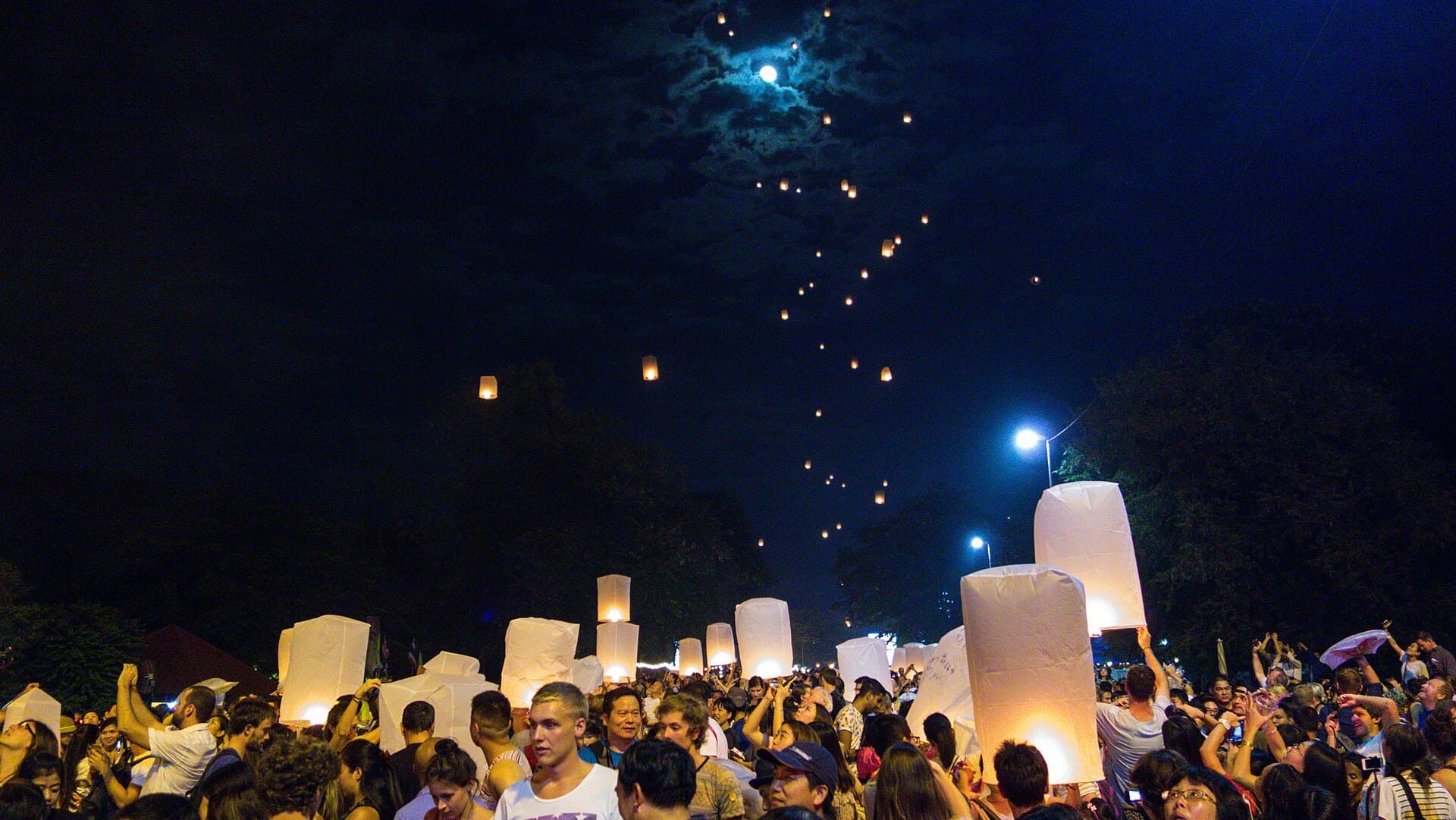 A crowd at the Yi Peng festival in Chiang Mai holding up glowing white paper sky lanterns, with a long trail of already-released lanterns climbing across the dark sky towards a bright moon behind thin cloud.