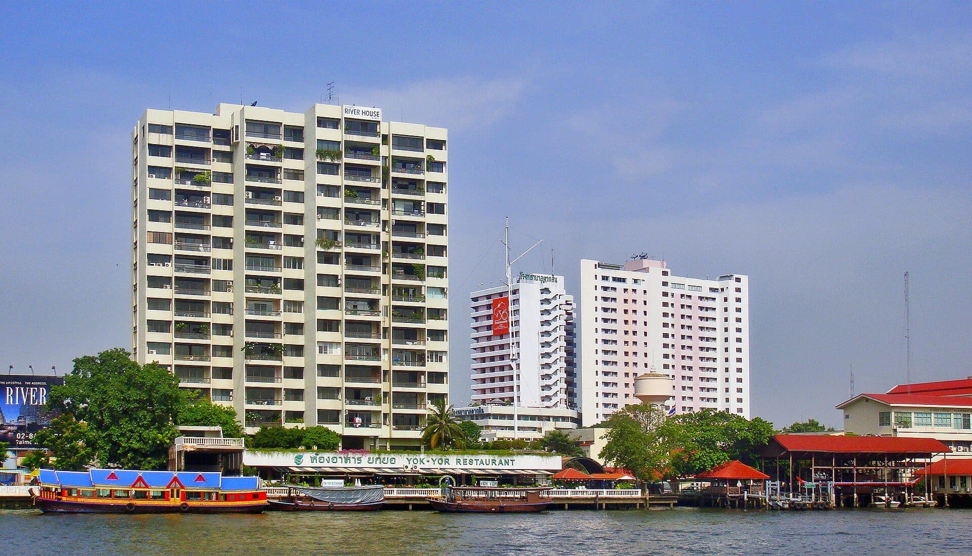 Mid-rise residential condominium blocks lining the bank of the Chao Phraya in Bangkok, their balconies facing the water above a riverside restaurant terrace and moored boats, with taller towers behind.